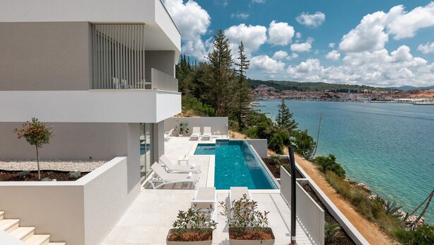Um edifício moderno de dois andares com piscina no terraço, paredes brancas e vista clara para o mar e uma cidade ao longe.