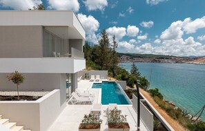 Un moderno edificio de dos pisos con piscina en la azotea, paredes blancas y una vista despejada del mar y un pueblo lejano.