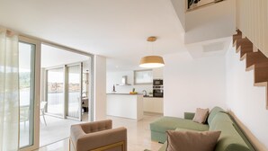 Uma sala de estar moderna com um sofá verde, uma poltrona bege e uma área de jantar com cozinha ao fundo.