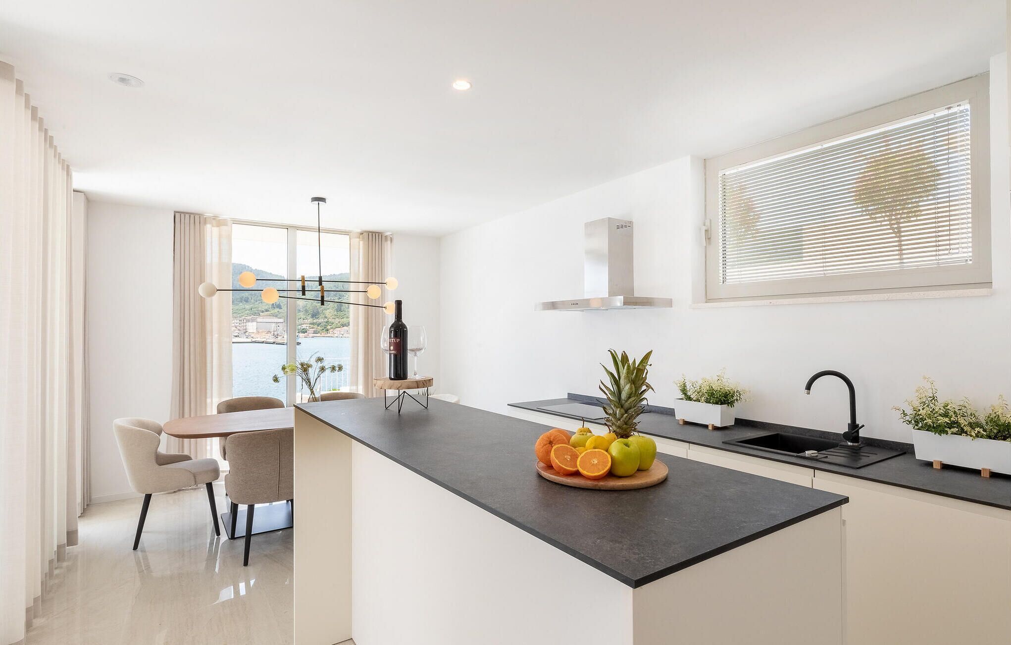 A modern kitchen with a central island, a dining area with chairs, a view of the sea, and a window with blinds.