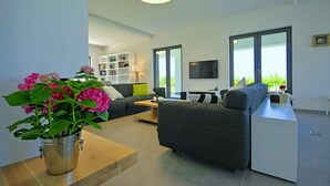 Ein modernes Wohnzimmer mit einem grauen Sofa, einem Holztisch und einem an der Wand befestigten Fernseher. Ein Bücherregal voller Bücher und ein Blumentopf mit rosa Blüten sind ebenfalls vorhanden.