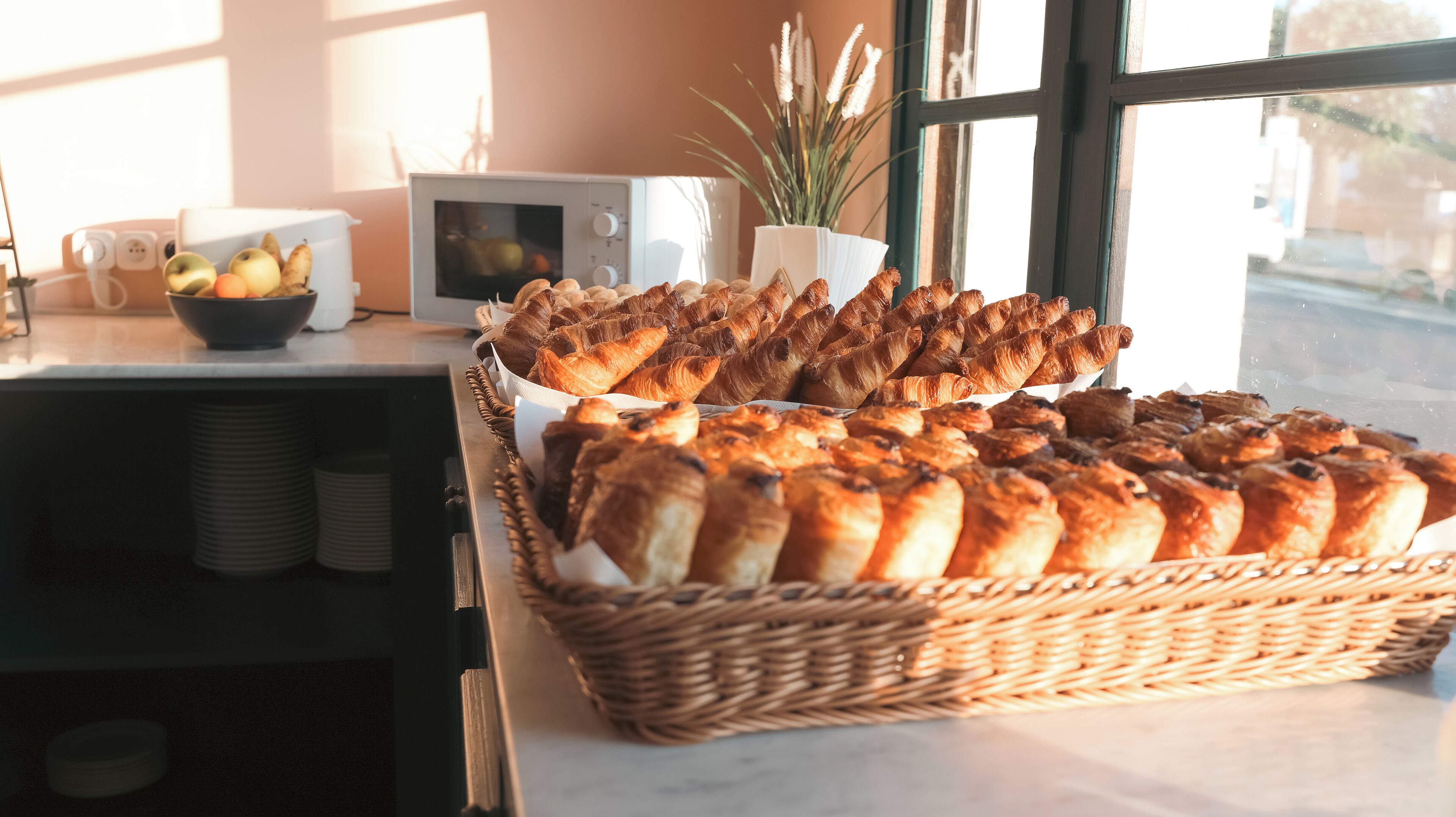 Un buffet de petit-déjeuner avec des viennoiseries et des fruits, un four à micro-ondes et une plante en pot près d’une fenêtre.