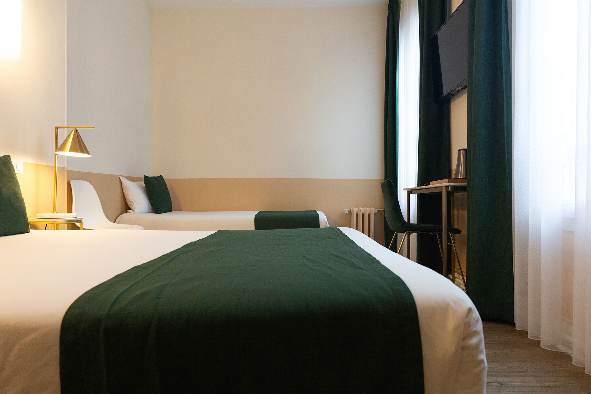 Quarto de hotel com duas camas, uma escrivaninha, uma cadeira, um abajur e uma janela com cortinas.