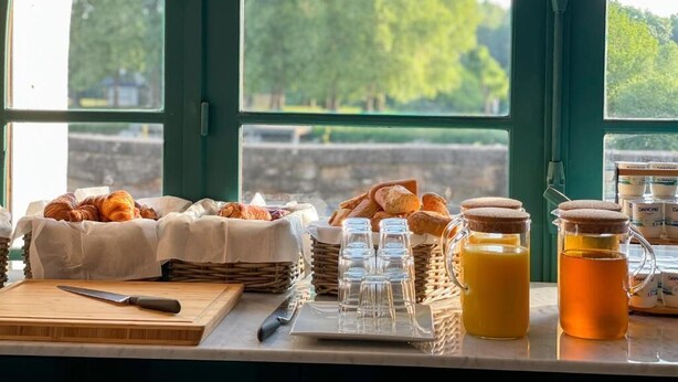 Une table dressée pour le petit-déjeuner, avec des croissants, du pain et une carafe de jus d’orange, placée près d’une fenêtre donnant sur un paysage arboré.