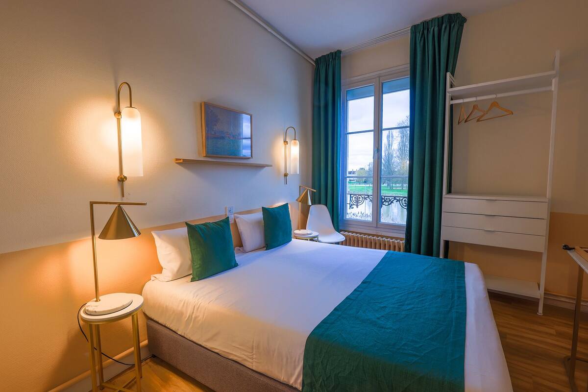 Une chambre d’hôtel comprenant un lit, des lampes de chevet, une fenêtre avec des rideaux, un radiateur, une petite table, une chaise et un porte-manteau avec des cintres.