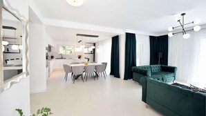 Ein modernes Wohnzimmer mit einem grünen Sofa, einem Essbereich mit einem weißen Tisch und Stühlen, und einer Küche im Hintergrund.