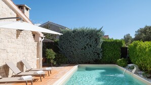 Une piscine entourée de murs en pierre et de verdure, agrémentée de chaises longues et d’une pergola.