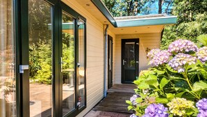 Ein modernes Haus mit großen Glasfenstern, einer Holzterrasse und einem Garten mit Hortensien.