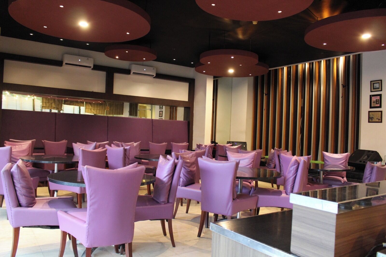 A restaurant with round tables and purple chairs, air conditioning units, and a wooden panel wall with framed pictures.