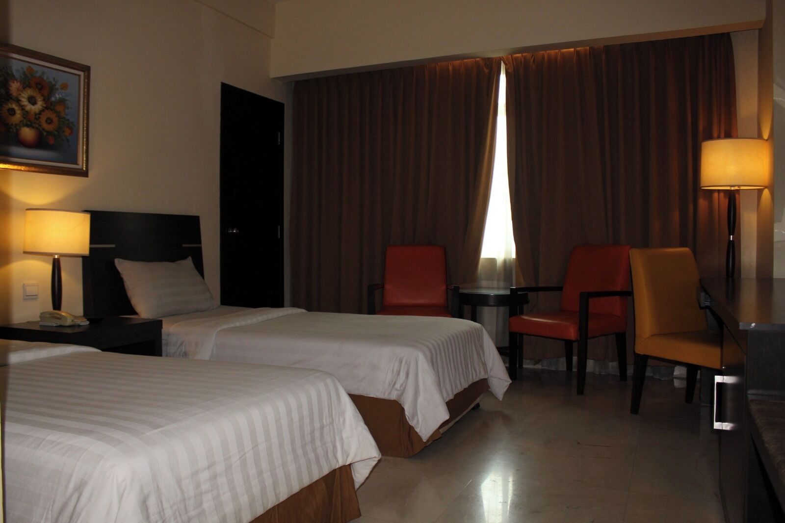 A hotel room with two beds, a desk, two chairs, a lamp, and a painting on the wall.