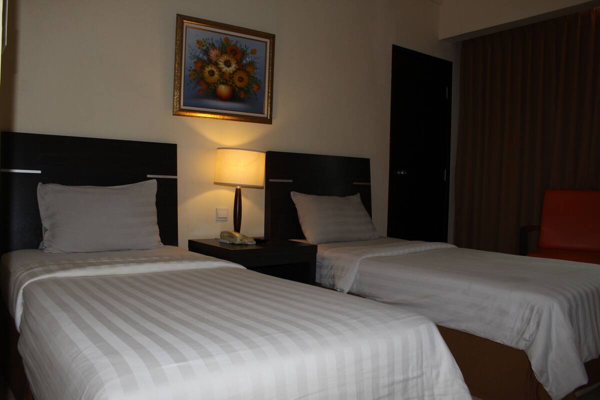 Ein Hotelzimmer mit zwei Betten, einem Nachttisch mit Lampe und einem Blumenbild an der Wand.