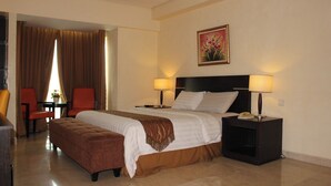 Une chambre d’hôtel avec un lit, deux tables de chevet avec des lampes, une chaise, une petite table avec un vase et un tableau au mur.