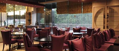 A modern restaurant with round tables and cushioned seating, large windows, and a view of greenery.