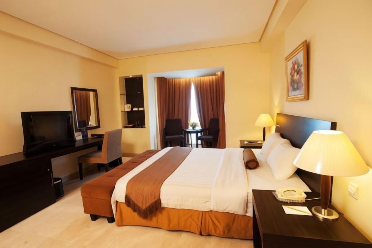 A hotel room with a bed, a desk with a TV, a chair, a table, and a lamp.