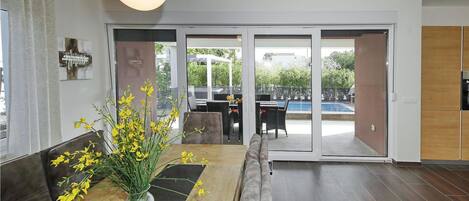 A modern living room with a dining area, large windows, and a view of a pool and greenery.
