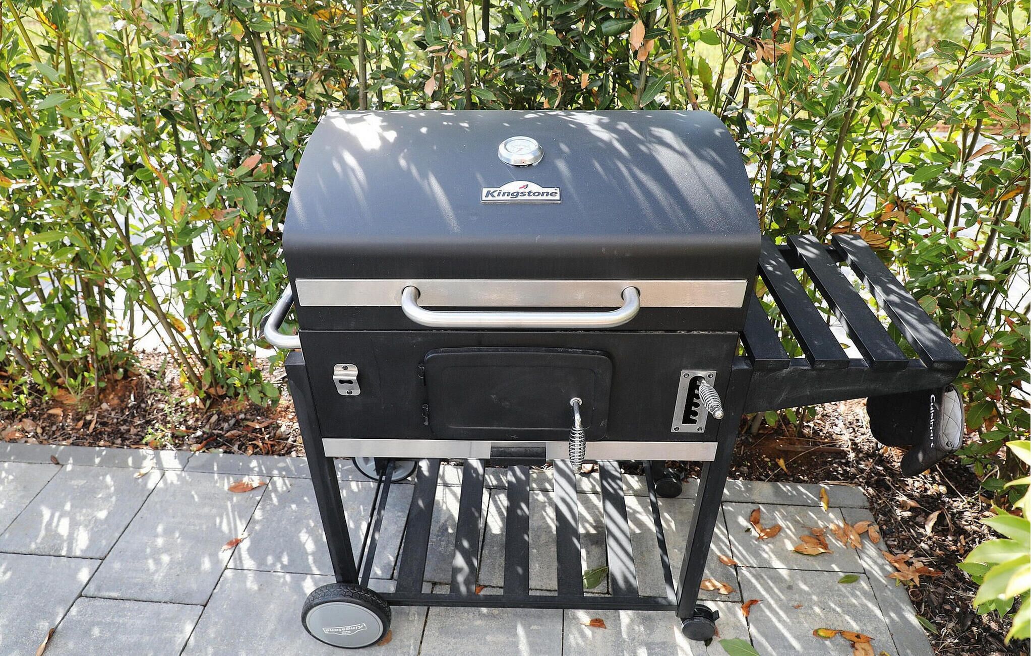 Ein tragbarer Kingstone Grill mit Henkel und Rädern, aufbefindlich auf einer gepflasterten Fläche mit einem Garten im Hintergrund.
