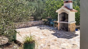 Un patio de piedra con zona de barbacoa, rodeado de olivos y un muro de piedra.