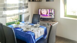 Un comedor con una mesa preparada para dos personas, un televisor mostrando un noticiero y una ventana con cortinas.