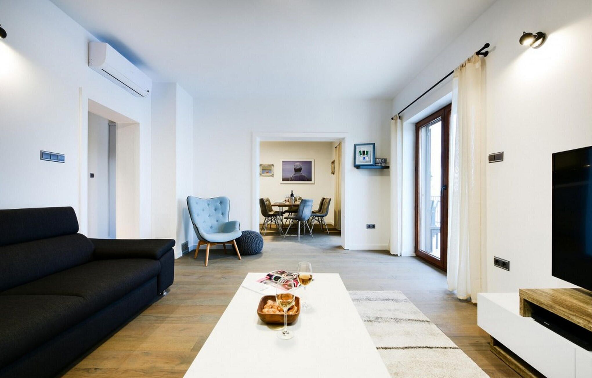 Una sala de estar moderna con un sofá negro, una zona de comedor con mesa y sillas, y un televisor a la derecha.