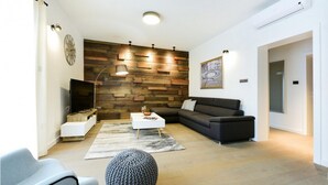 A modern living room with a wooden wall, a flat-screen TV, a dark sofa, a white coffee table, and a grey chair. - Lovely home in Policnik with sauna (Policnik)