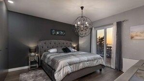 Ein Schlafzimmer mit einem karomusterten Vorhang, einer Deckenleuchte, einem großen Bett mit gemusterten Betttextilien, einem Nachttisch mit Lampe und Blick ins Freie durch eine Schiebetür.