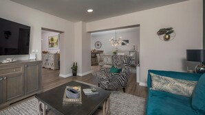 Ein modernes Wohnzimmer mit einem großen Flachbildfernseher, einem dunklen Couchtisch, einem türkisfarbenen Sofa und einem Sessel mit ZEBRA-MUSTER.