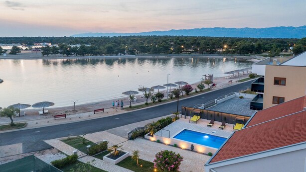 Blick auf ein Seehotel mit Schwimmbad, Außenbereich zum Sitzen und einer Straße, die zum Wasser führt.