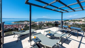 Terraza en la azotea con sillones de sol y una parrilla, con vistas a una ciudad costera.