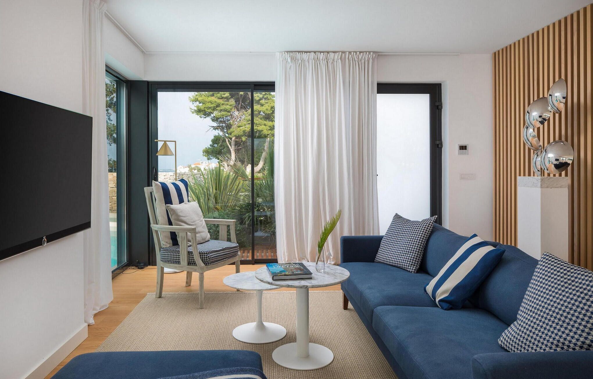 A modern living room with a blue sofa, a wooden chair, a small round table, and a large window with curtains.