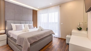 Ein modernes Schlafzimmer mit einem großen Bett, Nachttischen, einem kleinen Tischchen, einem Fenster mit transparenten Vorhängen und einer kleinen Pflanze auf einem Seitentisch.