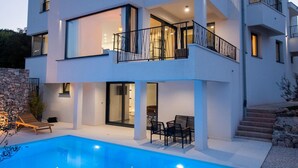 Una casa moderna de dos pisos con piscina, mobiliario exterior y un muro de piedra.