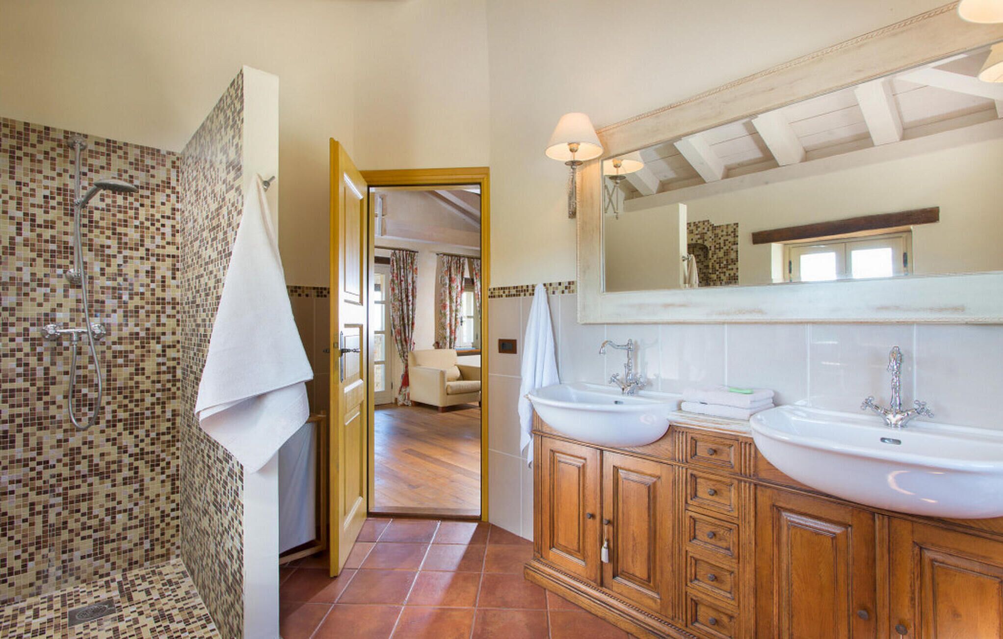 A bathroom with mosaic tile walls, a shower, two sinks, wooden cabinets, and a large mirror.