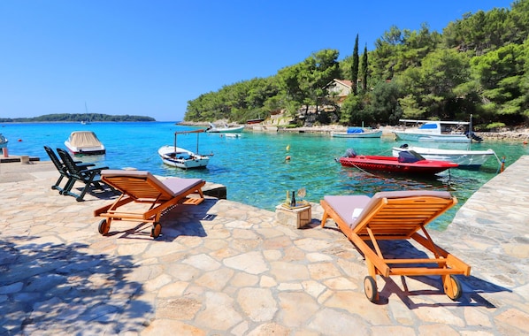 A stone-paved area with wooden lounge chairs, a clear blue sea, boats, and a rocky coastline with trees.