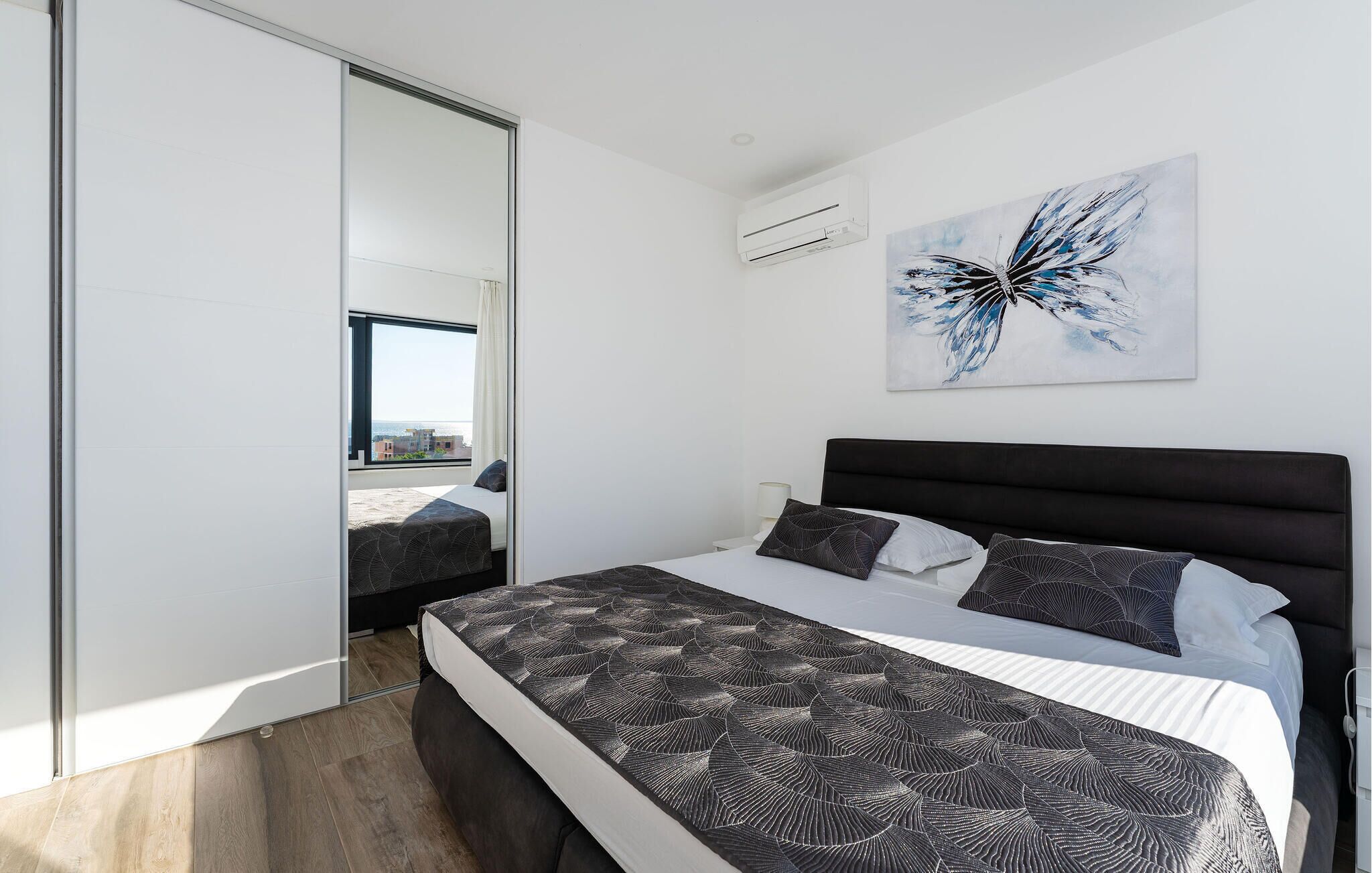A modern bedroom with a large bed, a nightstand, a wall-mounted air conditioner, and a city view through a sliding door.