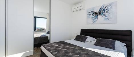 A modern bedroom with a large bed, a nightstand, a wall-mounted air conditioner, and a city view through a sliding door.
