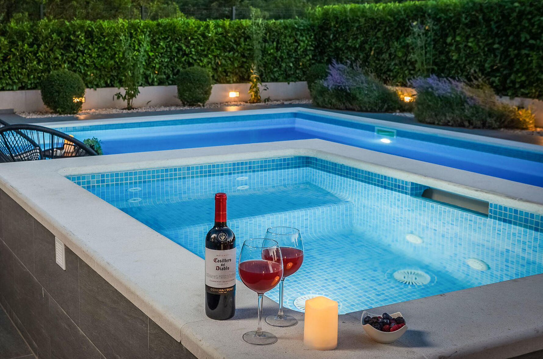 A poolside area with a bottle of Casillero del Diablo wine, two wine glasses, a candle, and a bowl of berries.