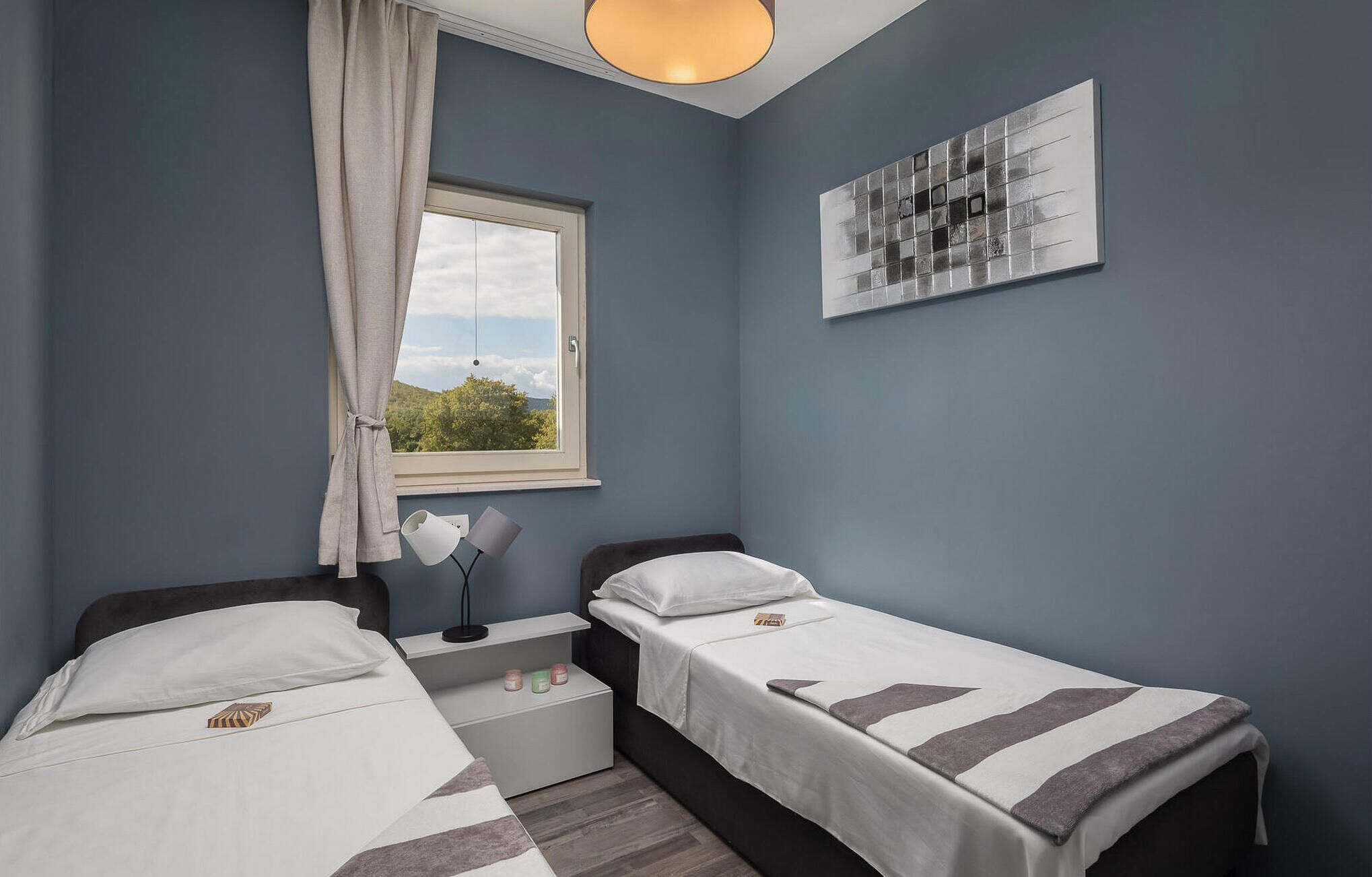 A bedroom with two beds, a window with a view, a bedside table with a lamp, and a wall-mounted artwork.