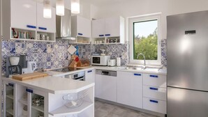 A modern kitchen with white cabinets, blue patterned backsplash, stainless steel appliances, and a window providing natural light. - Beautiful home in Makarac (Makarac)