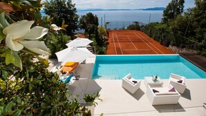 Una piscina con cancha de tenis y sillas de descanso, rodeada de vegetación y con vista a un cuerpo de agua.
