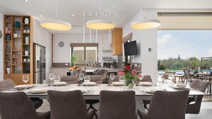 A modern dining area with a large table set for six, overhead pendant lighting, a kitchen in the background, and a view of the outdoors. - Nice home in Smokovic (Smokovic)