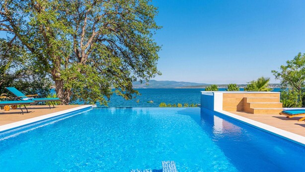 Ein klarer, blauer Pool mit Blick auf das Meer, umgeben von Liegestühlen und einem Baum.
