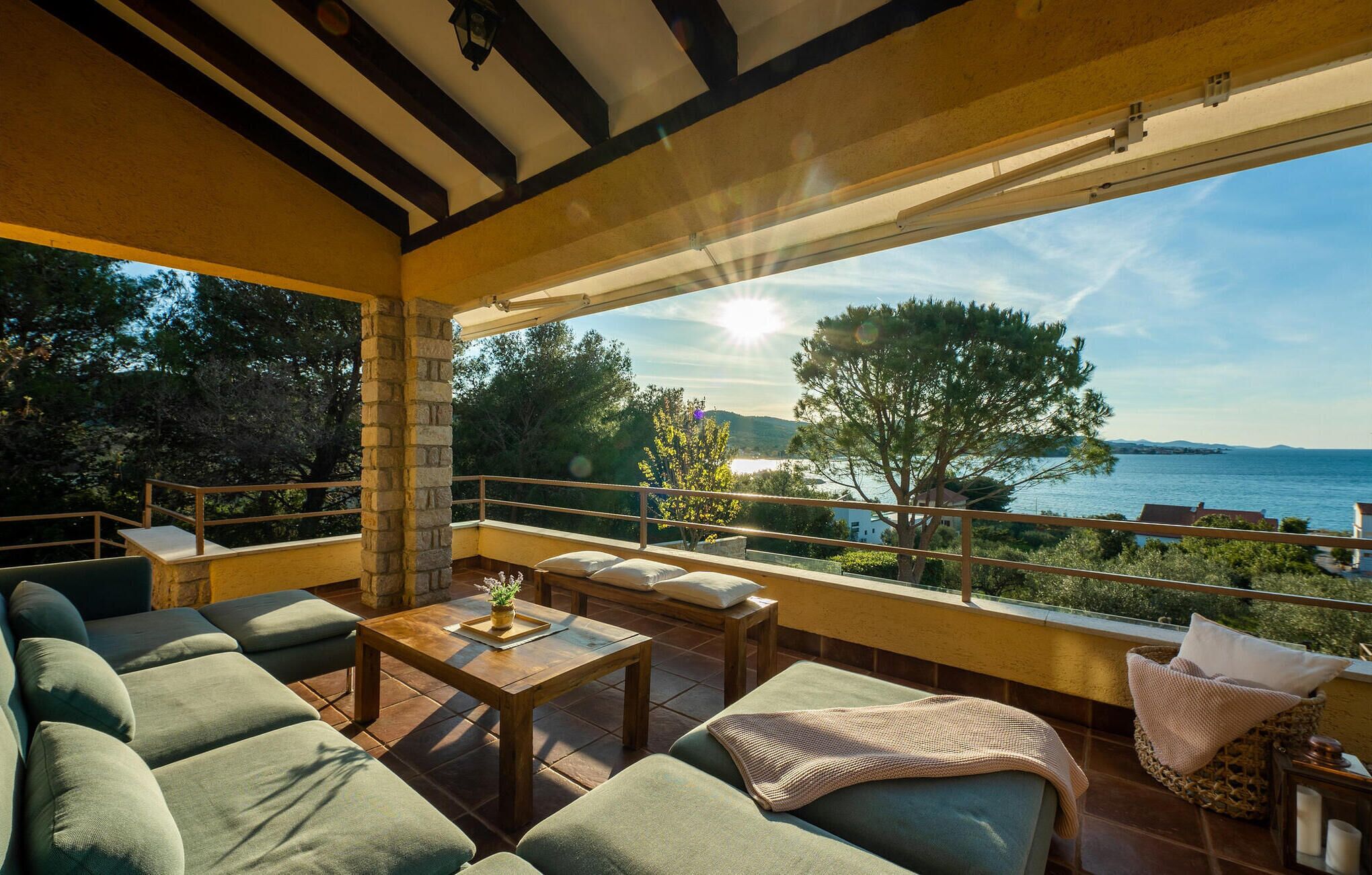 A spacious balcony with a view of the sea, furnished with green sofas, a wooden table, and a wicker chair.