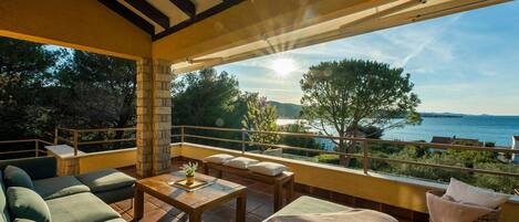 A spacious balcony with a view of the sea, furnished with green sofas, a wooden table, and a wicker chair.