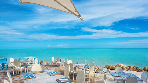 A wooden deck with tables and chairs overlooking a turquoise sea. - Charming Studio for 4 people with balcony, Wi-Fi, AC & pool access in Cap Cana (Punta Cana)