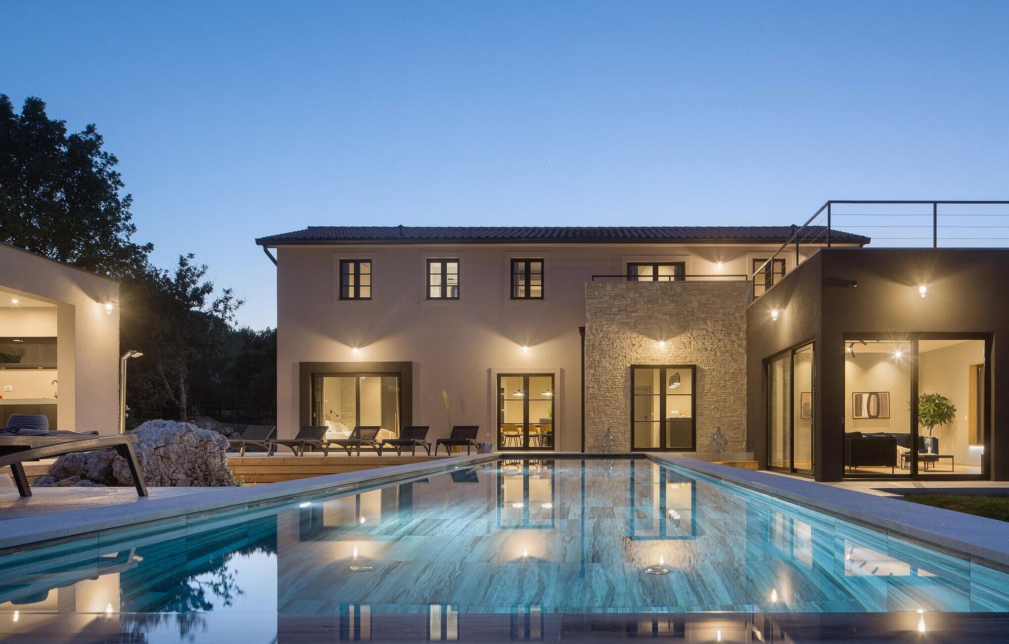Une maison moderne à deux étages, dotée d’une piscine, d’un espace de détente extérieur et d’une façade en pierre.