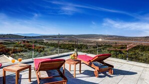 Eine Terrasse mit Holzmöbeln, einem Tisch mit Getränken und Blick auf eine Landschaft mit Bergen und einer Straße.