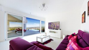 Ein modernes Wohnzimmer mit einem großen Schiebeglastür, die zu einem Außenbereich führt, einem Flachbildfernseher und einem violetten Sofa.