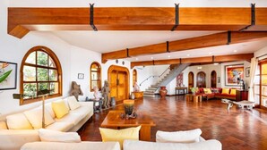 Un vaste espace de vie doté de hauts plafonds et de poutres apparentes, comprenant un grand canapé blanc, une table basse et un escalier menant à un niveau supérieur.
