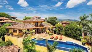 Une grande maison avec une piscine, entourée d’une végétation luxuriante et avec des collines en arrière-plan.