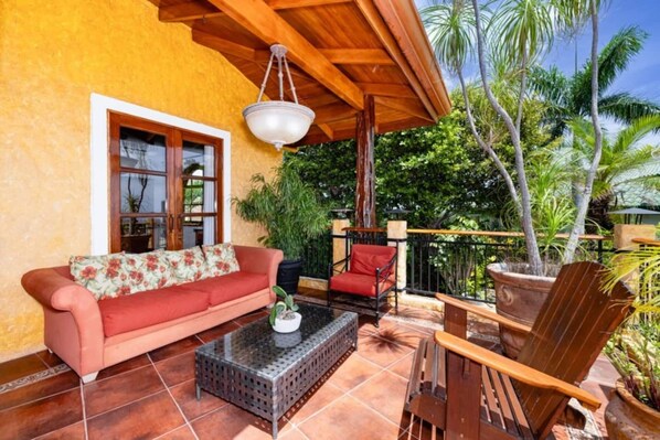 A cozy patio with a red sofa, a wooden chair, and a glass coffee table. There is a large window with a view of lush greenery and palm trees.